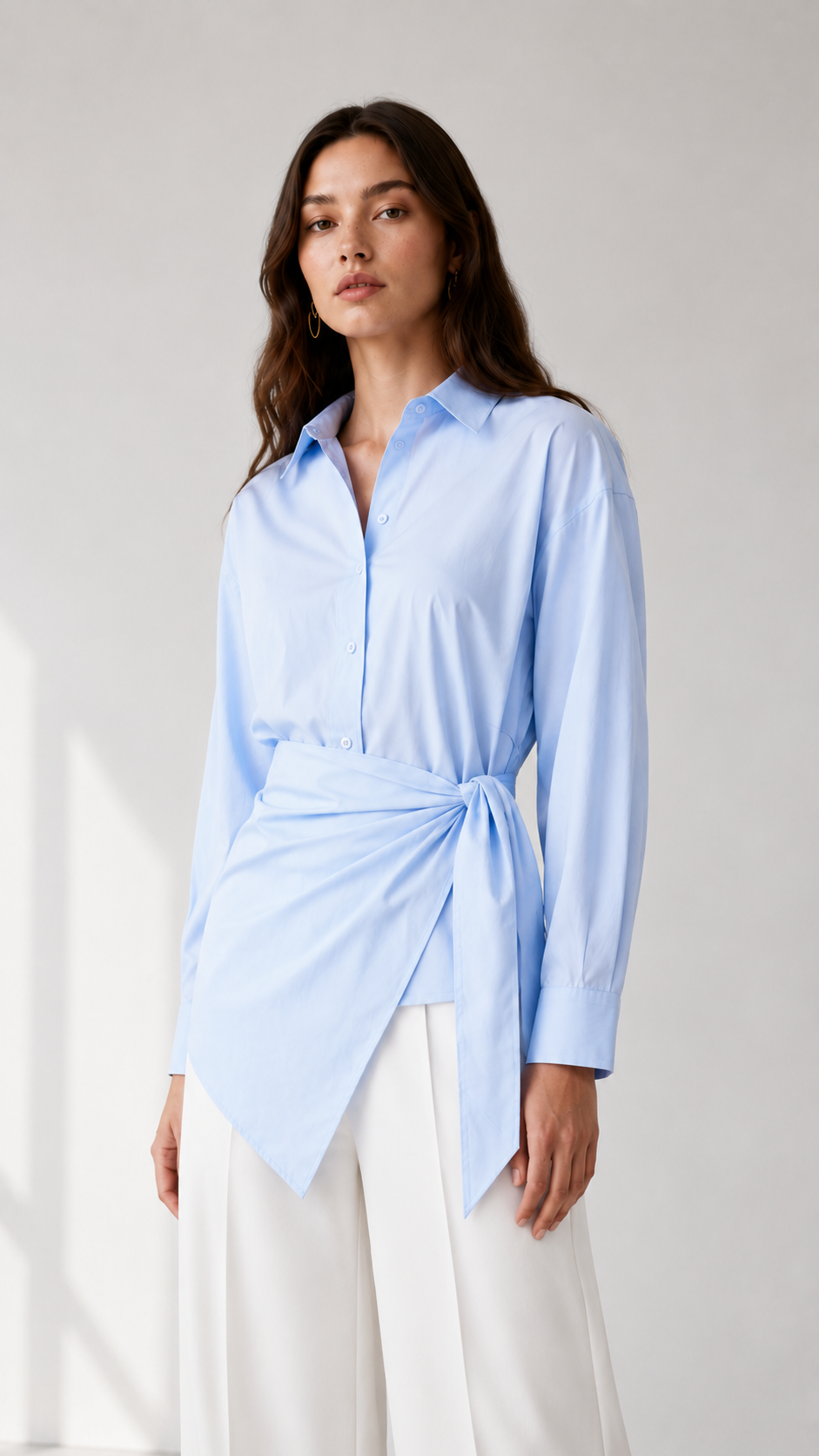 Wrap-front button-down shirt in pastel blue, yellow, and pink. It features a classic pointed collar, concealed-style front button placket, long sleeves with wide cuffs, and a waist tie detail that gathers at the side for a flattering draped silhouette. The asymmetric wrap hem adds a polished yet feminine touch, ideal for workwear, brunch, or elevated everyday styling.